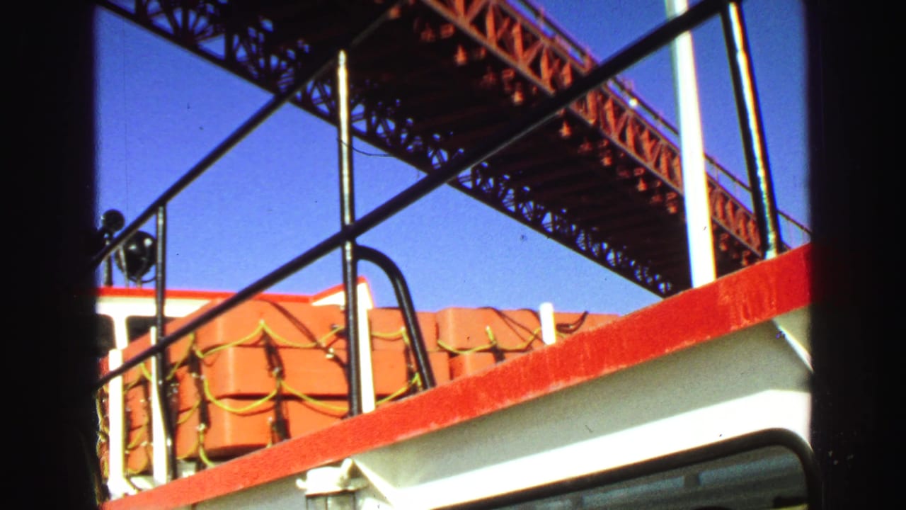 1968 Vancouver boat view with red railing, orange life vests, and the underside of a steel truss bridge against blue sky.