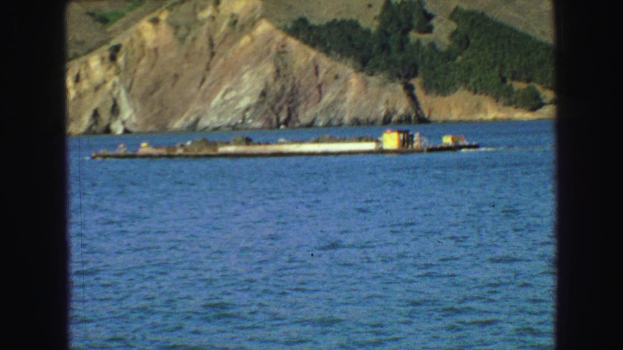 Long barge on blue water with a rocky hillside behind it.