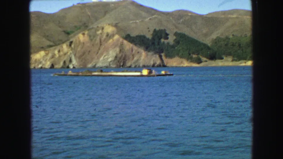 1968 Vancouver boat-window view of a barge on blue water, backed by hills and scattered shoreline trees.