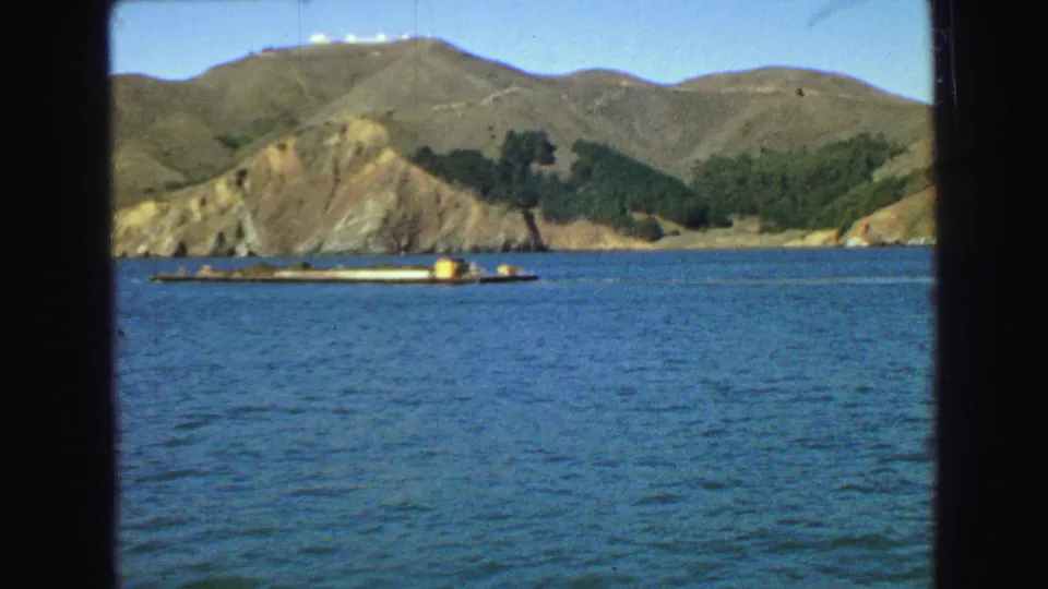 1968 Vancouver harbor view from a boat, with a low barge on the water and rounded hills beyond the shoreline.