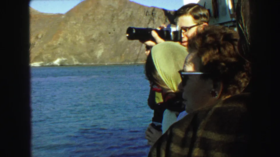 1968 Vancouver boat passengers in sunglasses and headscarf look across blue water while a man photographs the mountainous coast.