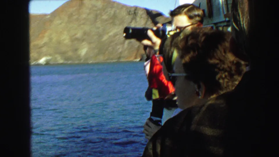 1968 home-movie frame of boat passengers sightseeing near the Golden Gate Bridge, with a man aiming a camera toward the bay.