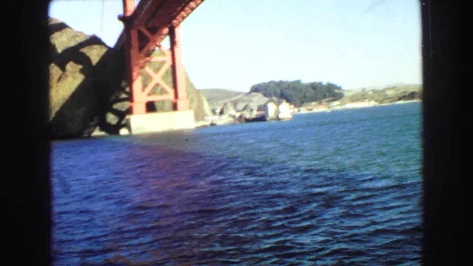 View from a boat toward the Golden Gate Bridge tower, blue water, rocky shore, and distant buildings in daylight.