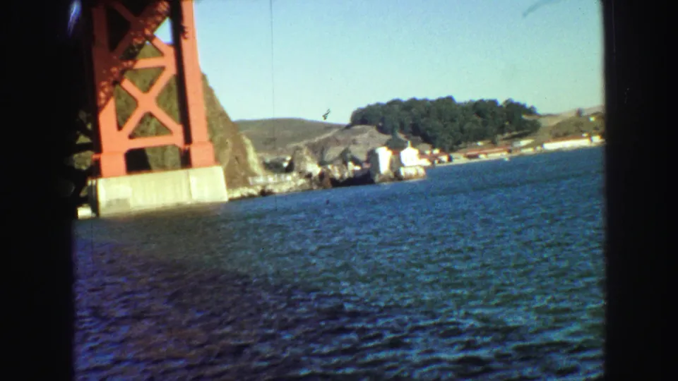 Boat-level view of the Golden Gate Bridge tower with bay water, rocky shoreline, low hills, and a cluster of trees in the distance.
