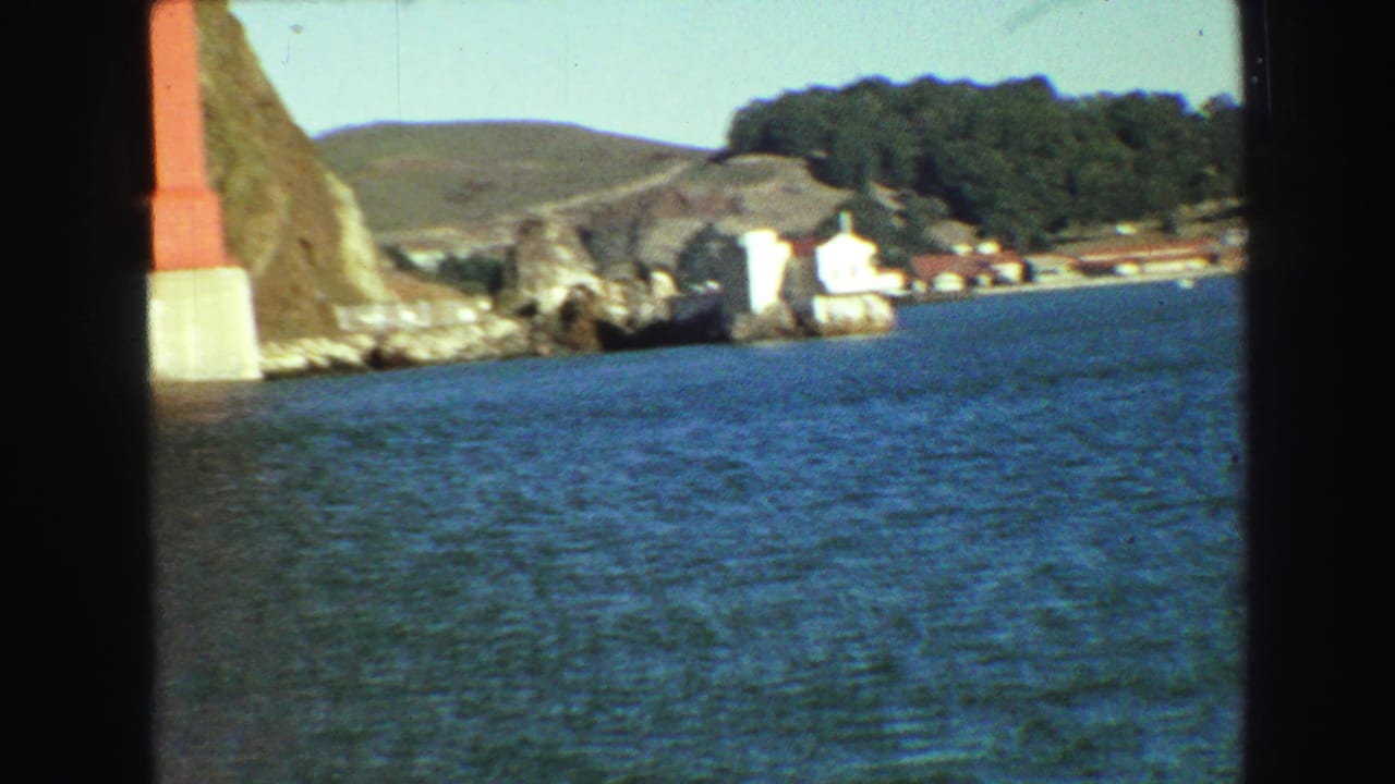 Bay water with rocky shoreline, white buildings, hills, and a red bridge support at left.