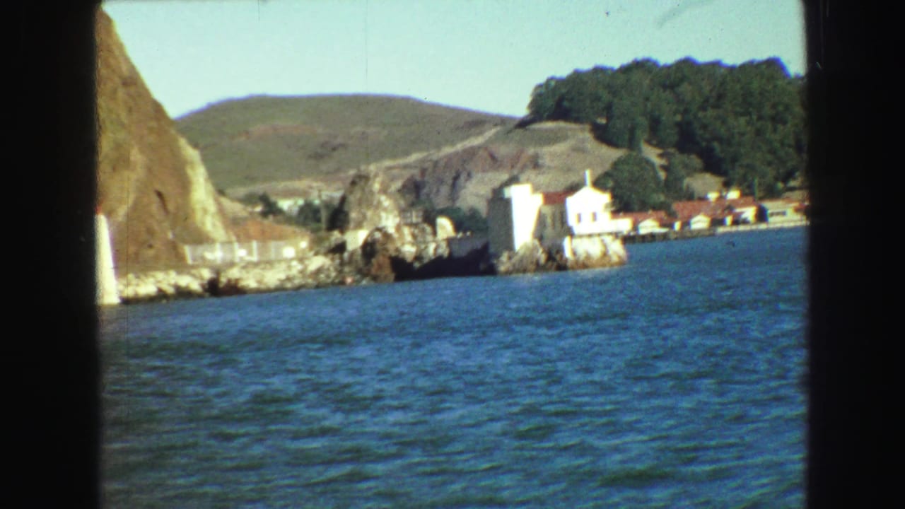 Coastal water with rocky outcrops, white buildings, low hills, and trees in the distance.