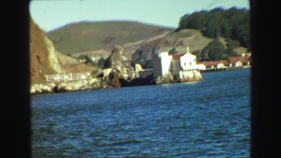 1968 8mm travel view across bay water toward rocky shoreline, white buildings, wooded hills, and pale sky.