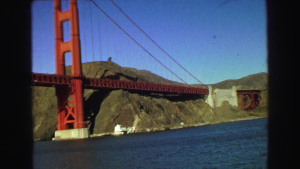 1968 8mm frame showing a red suspension bridge tower, bridge deck, cables, blue water, and coastal hills.