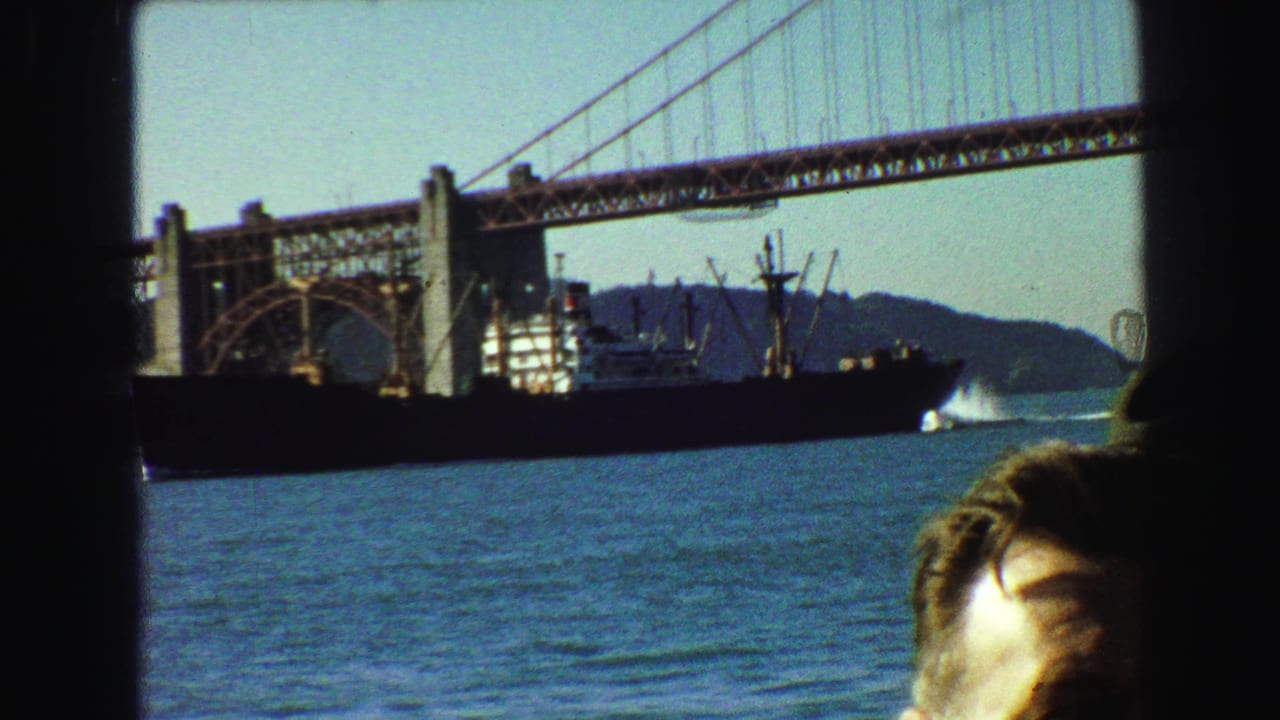 1968 8mm frame of Golden Gate Bridge seen from a boat, with a large ship on the water and a cropped passenger in front.
