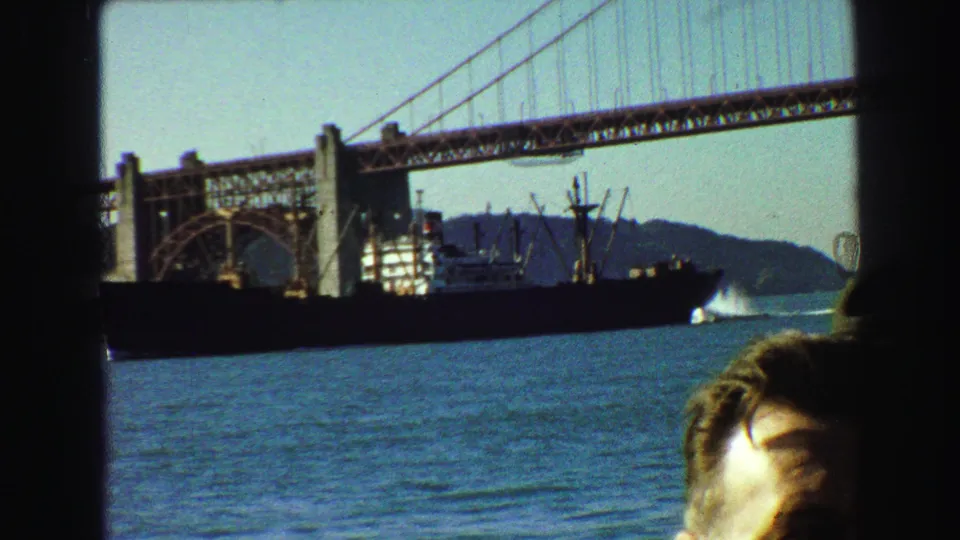 1968 8mm frame of Golden Gate Bridge seen from a boat, with a large ship on the water and a cropped passenger in front.