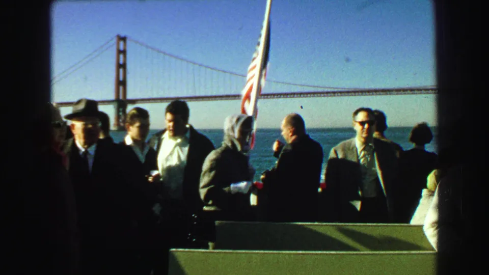 1968 8mm frame of adult passengers on a boat deck near an American flag, with Golden Gate Bridge in the background.