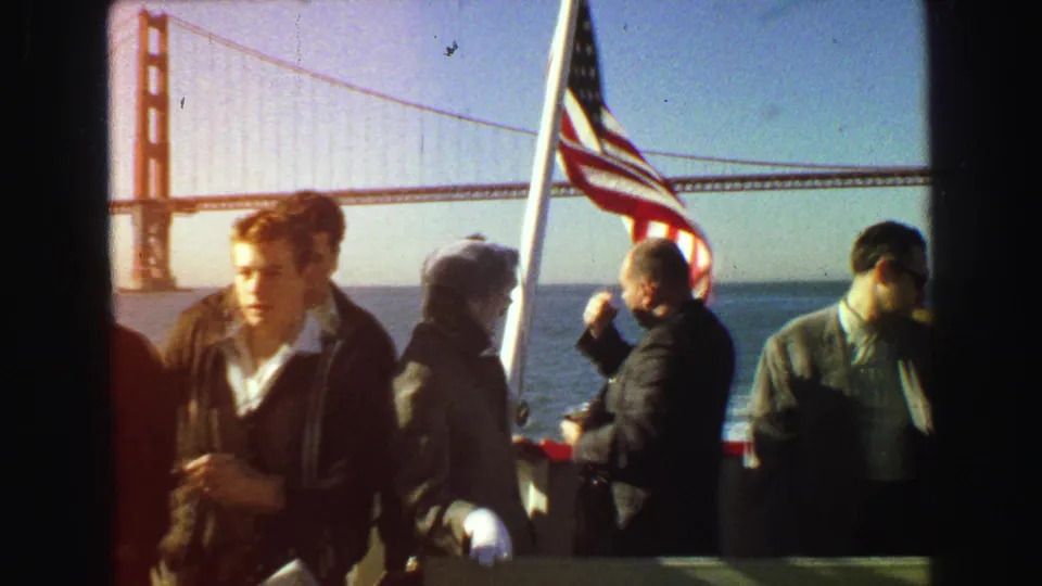 1968 8mm frame showing passengers in period clothing on a boat, with a U.S. flag and Golden Gate Bridge behind them.