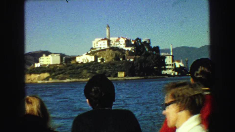 1968 home-movie frame with a foreground passenger in sunglasses and other tourists facing Alcatraz across the bay.