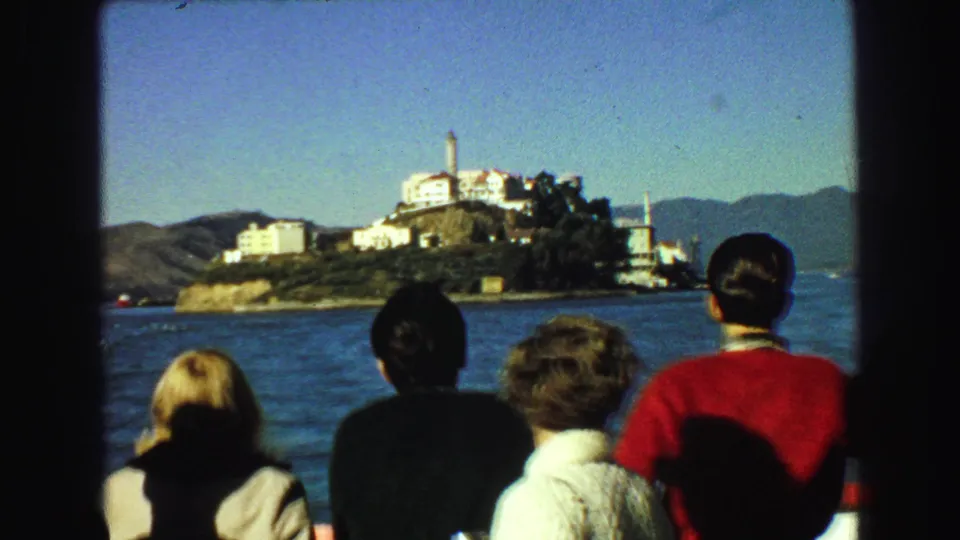 Tourists aboard a sightseeing boat face Alcatraz Island, with bay water, white prison buildings, and hills visible in the distance.
