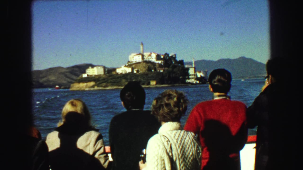 Tourists on a boat look toward Alcatraz Island across blue bay water.