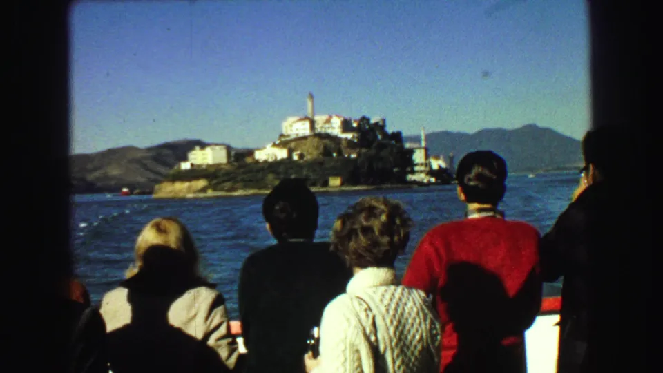 1968 8mm travel frame showing boat passengers viewed from behind as they face Alcatraz Island under a clear sky.