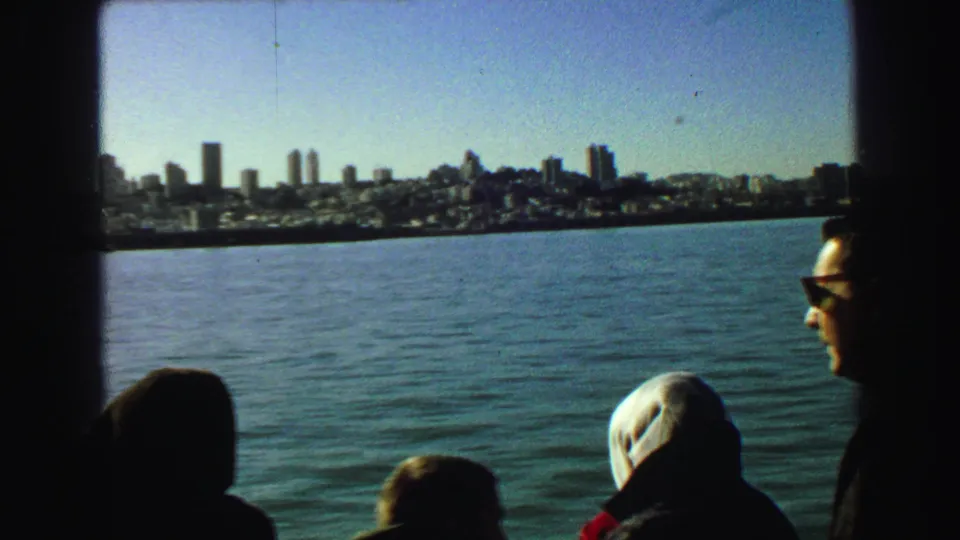 1968 8mm boat-tour view with seated passengers in the foreground and the San Francisco waterfront beyond open water.