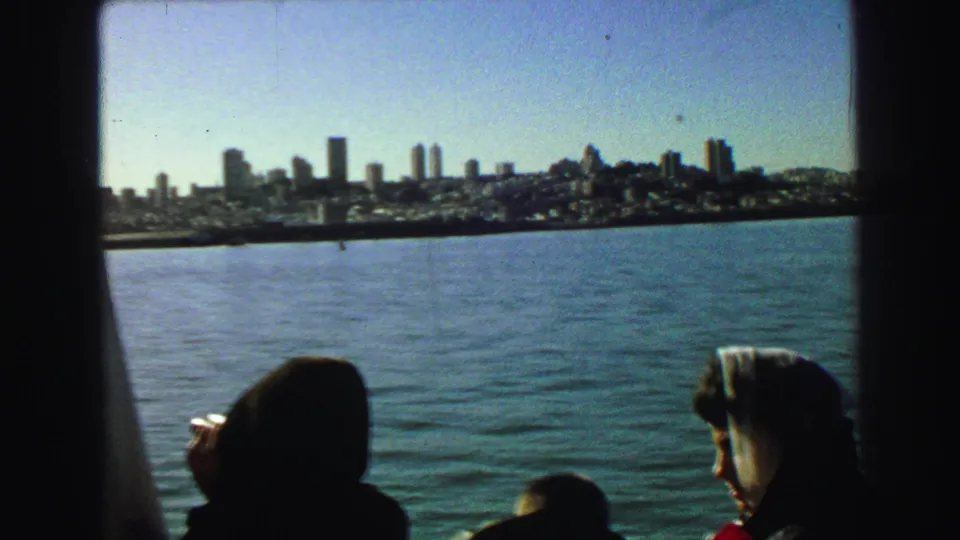 1968 8mm frame with silhouetted boat passengers in the foreground and San Francisco's waterfront skyline in the distance.