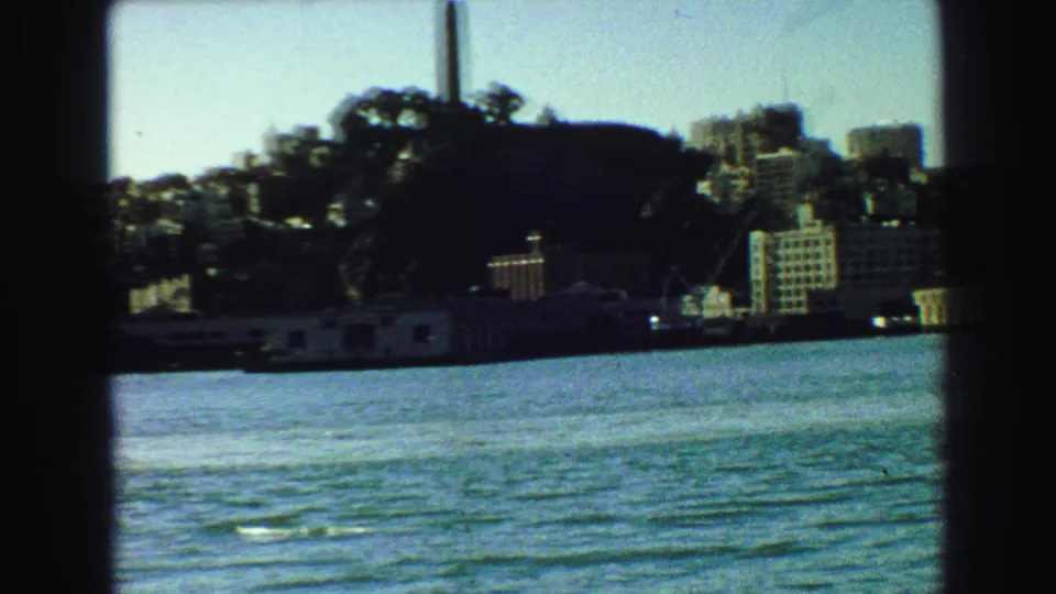 1968 Vancouver shoreline view with a tall smokestack, hillside trees, rippling water, and waterfront buildings framed by the boat window.