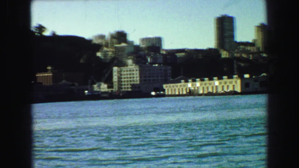 1968 Vancouver waterfront view through a boat window, showing water, shoreline buildings, hillside trees, and a tall tower in the distance.
