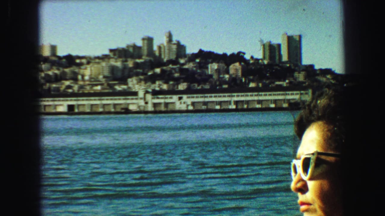 1968 Vancouver home-movie frame showing a sunglassed passenger by a dark window frame, with waterfront buildings, skyline, and water outside.