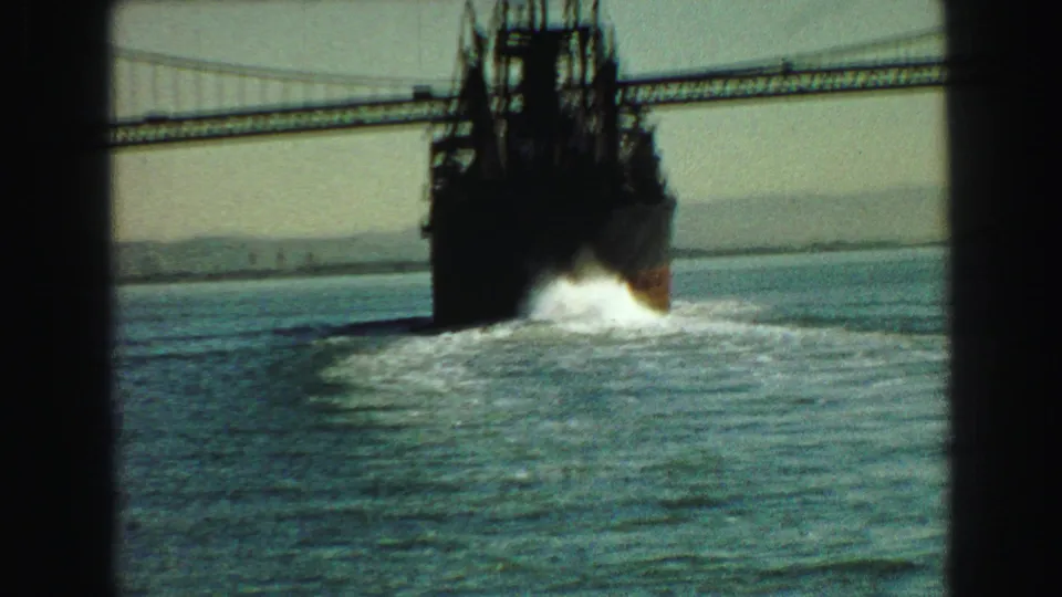1968 Vancouver harbor view of a large ferry or vessel passing beneath a suspension bridge, framed by dark window bars and water wake.