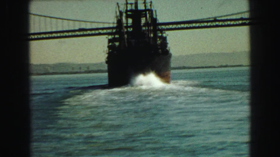 1968 Vancouver harbor footage frame with a ferry moving away below a suspension bridge, white wake trailing across the water.