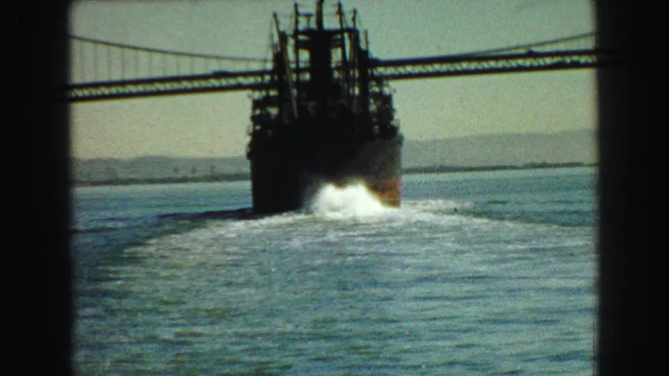 1968 Vancouver-area home-movie frame showing a large cargo ship moving under a suspension bridge with wake on the water.