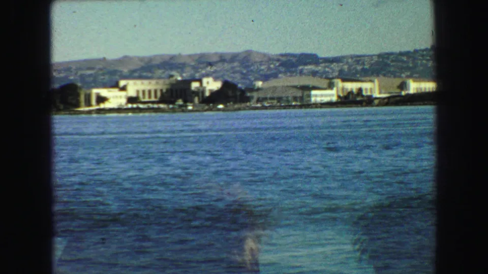 1968 Vancouver-area harbor frame with a small tugboat on open water and distant shoreline buildings below hills.