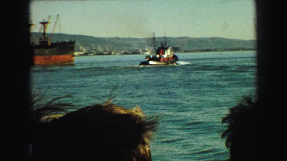 1968 Vancouver harbor view from a passenger area, with silhouetted heads watching a tugboat near a cargo ship on the water.