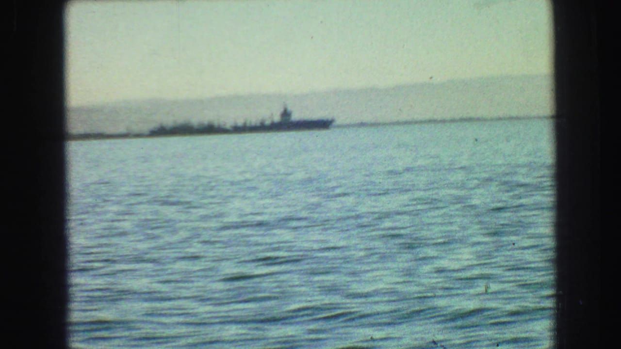 1968 Vancouver harbor view from 8mm footage, showing a distant battleship-like vessel across rippled water beneath a hazy shoreline.