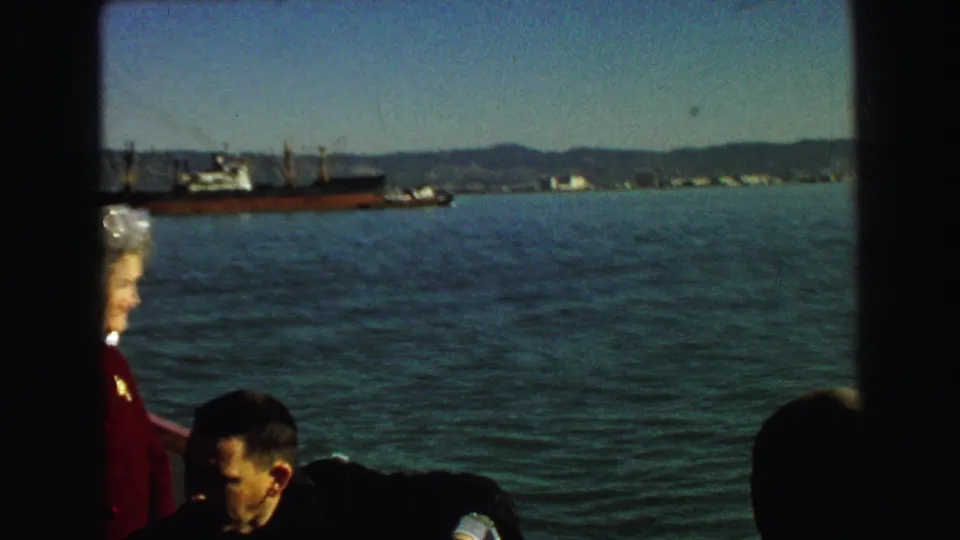 From a moving vessel in 1968 Vancouver, passengers sit and stand near the rail as cargo ships and shoreline hills appear across the harbor.