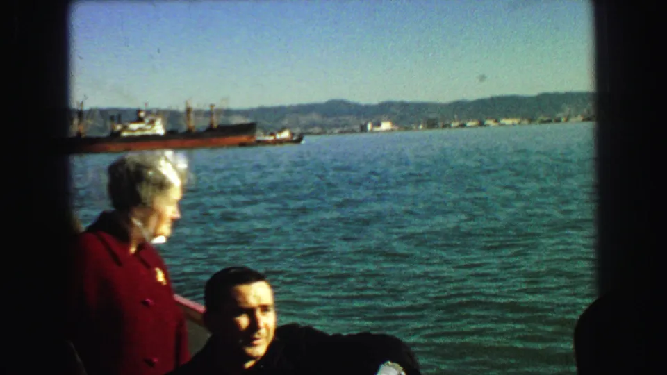 1968 Vancouver harbor footage shows a man and woman aboard a vessel, with blue-green water, cargo ships, and low shoreline hills behind them.