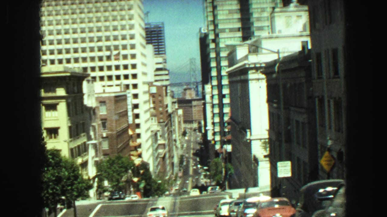 1968 Vancouver downhill street view with modern towers, older facades, traffic, and blue water beyond.
