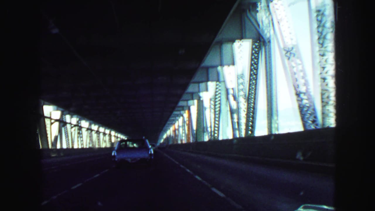 1968 Vancouver home-movie view from a car inside a dark steel-truss bridge, following a white sedan in the lane ahead.