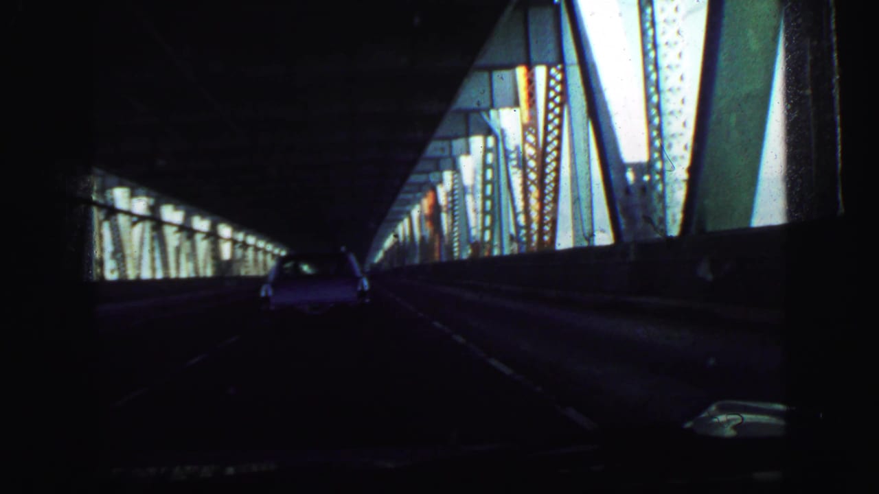 Dark 8mm passenger viewpoint inside the steel-truss bridge, showing the road, side beams, and a sedan ahead in traffic.