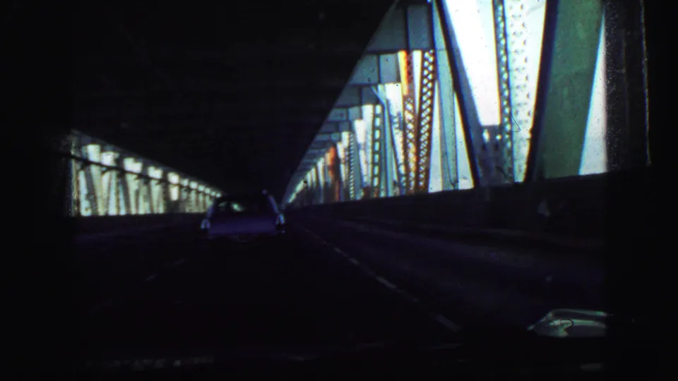 Dark 8mm passenger viewpoint inside the steel-truss bridge, showing the road, side beams, and a sedan ahead in traffic.