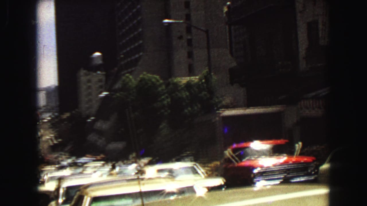 Urban street with parked cars, a red car, trees, tall buildings, and dark film gate edges.