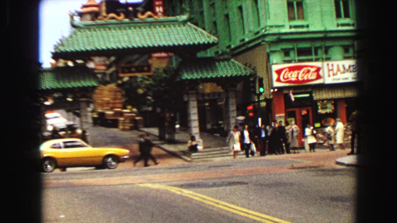 1968 Vancouver street view with a yellow car passing pedestrians near green tiled roofing, storefront signs, and a Coca-Cola sign.