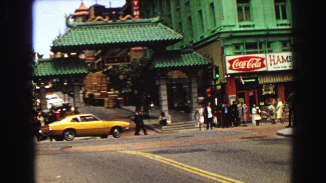 Chinese-style gate on a city street with pedestrians, storefront signs, and a yellow car passing.
