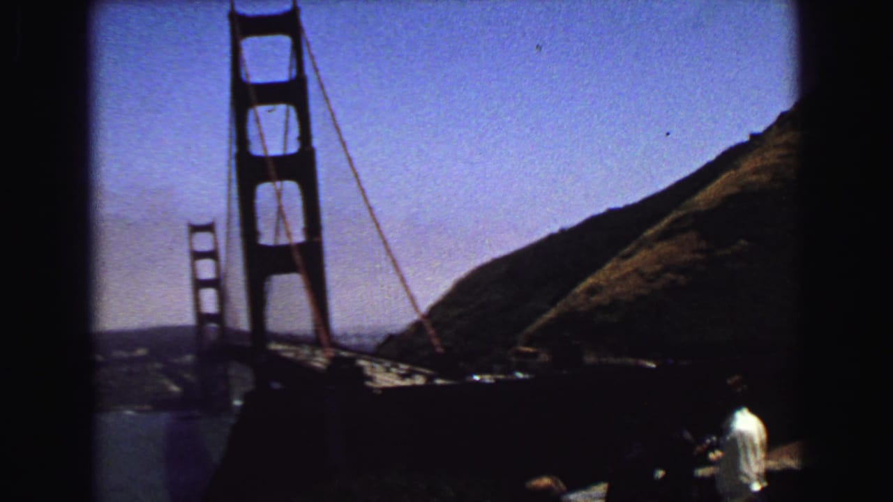 Visitor-shot 8mm view of the Golden Gate Bridge tower, adjacent hillside, bay water, and a person standing near the overlook edge.