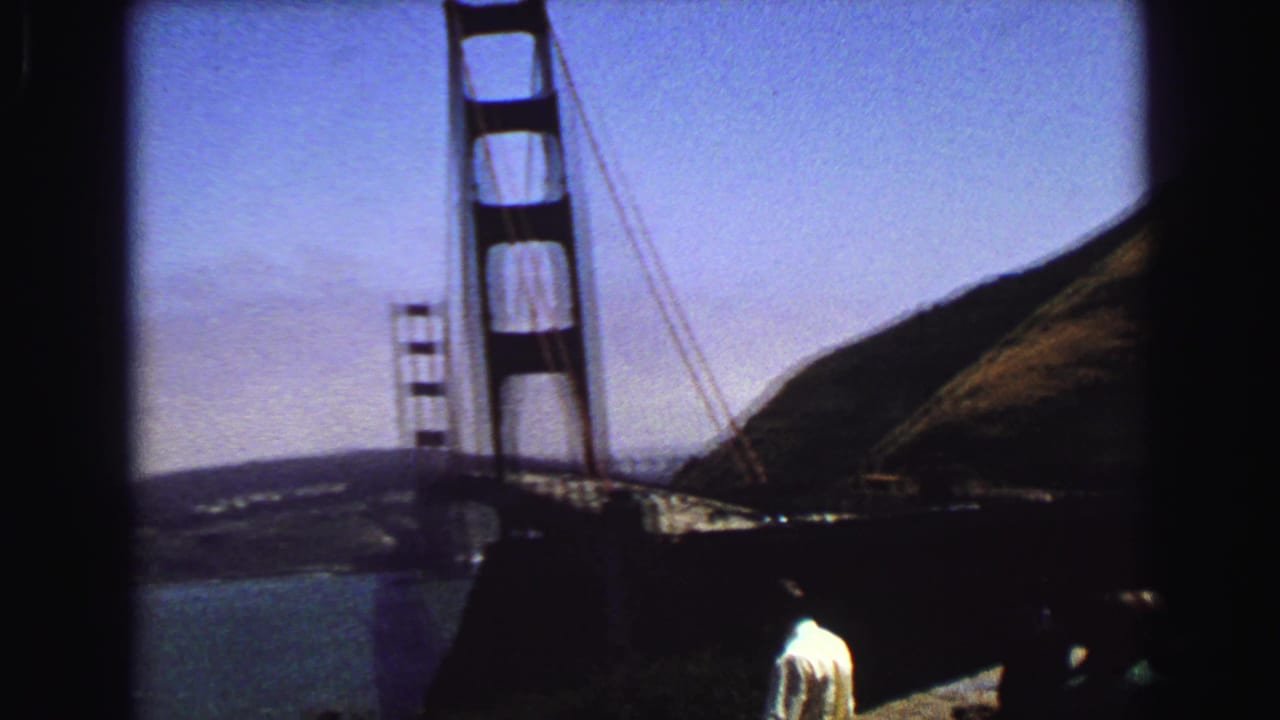 Handheld 8mm view of Golden Gate Bridge towers, bay water, hillside, and visitors partly visible near the overlook foreground.