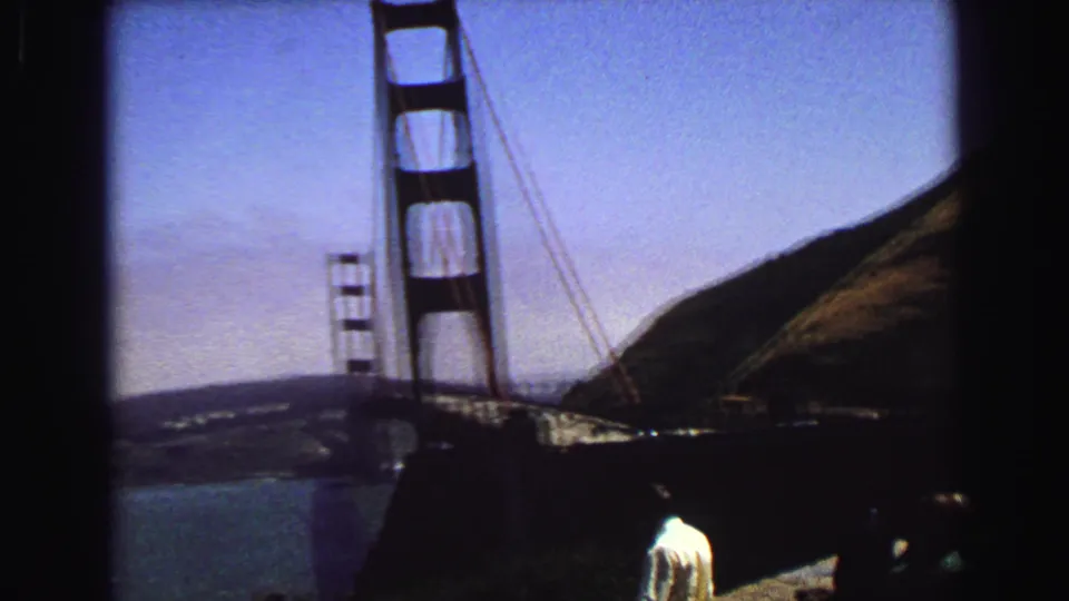Handheld 8mm view of Golden Gate Bridge towers, bay water, hillside, and visitors partly visible near the overlook foreground.