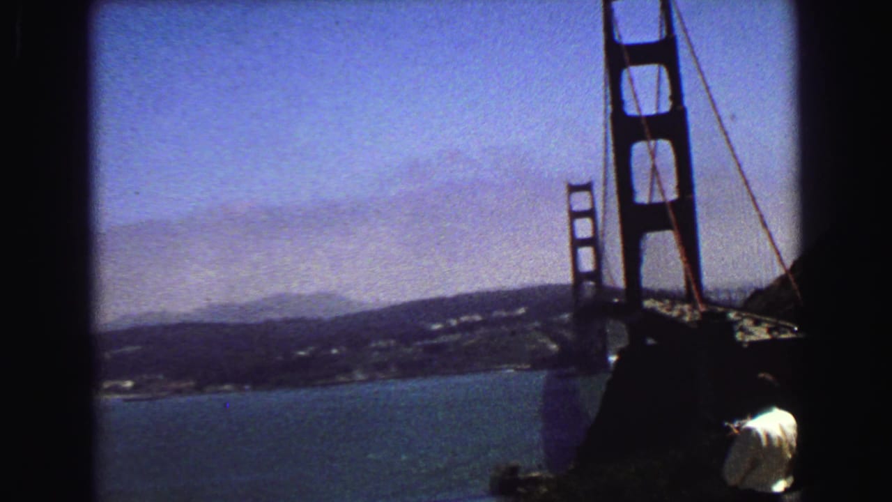 8mm travel view of the Golden Gate Bridge tower with bay and distant shoreline, framed by hillside and a visitor in a white shirt.