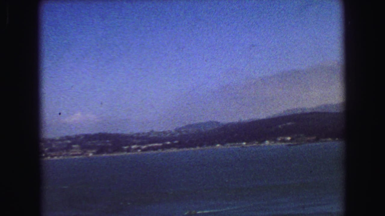 1968 8mm frame looking across blue water toward a distant coastal city shoreline and low hills.