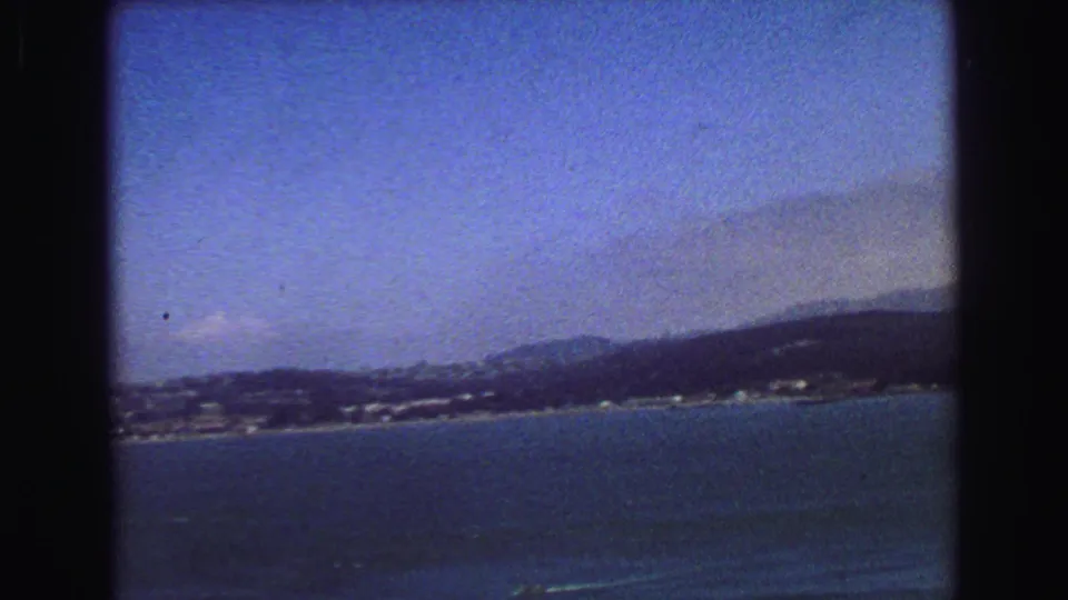 1968 8mm frame looking across blue water toward a distant coastal city shoreline and low hills.