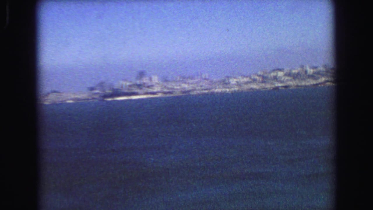 Blue-tinted 8mm frame of Vancouver's distant shoreline and city buildings across a wide water foreground.