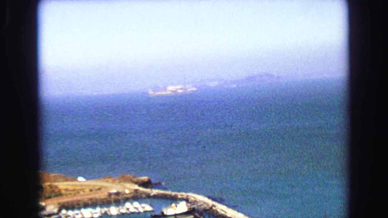 1968 Vancouver-area harbor view from above, showing moored boats, a breakwater pier, blue water, and hazy distant land.