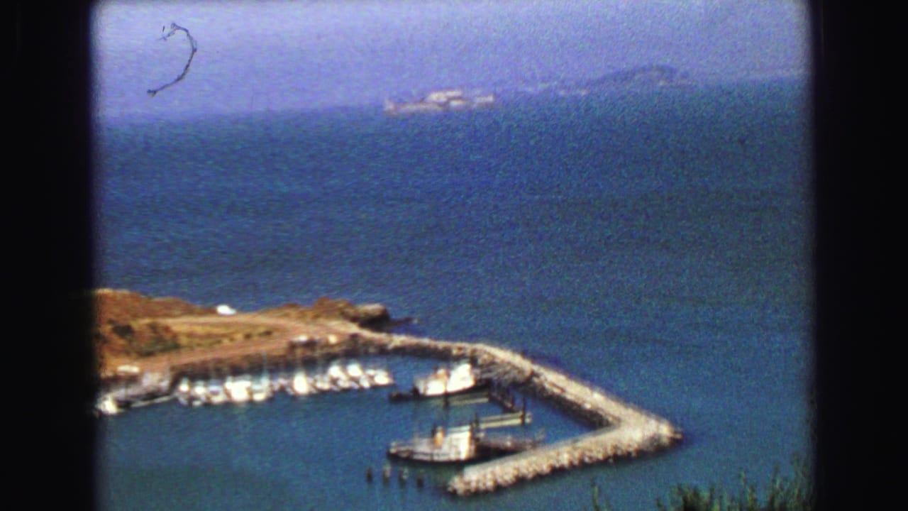 1968 Vancouver-area 8mm frame of a harbor basin, docked boats, stone pier, rocky shore, and distant hazy coastline.