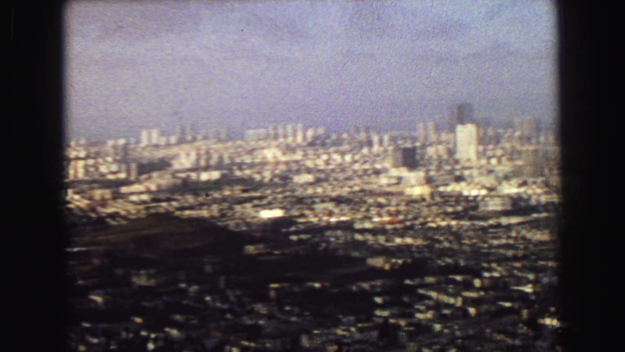 Distant cityscape seen through a dark window-like frame with towers and dense blocks under haze.